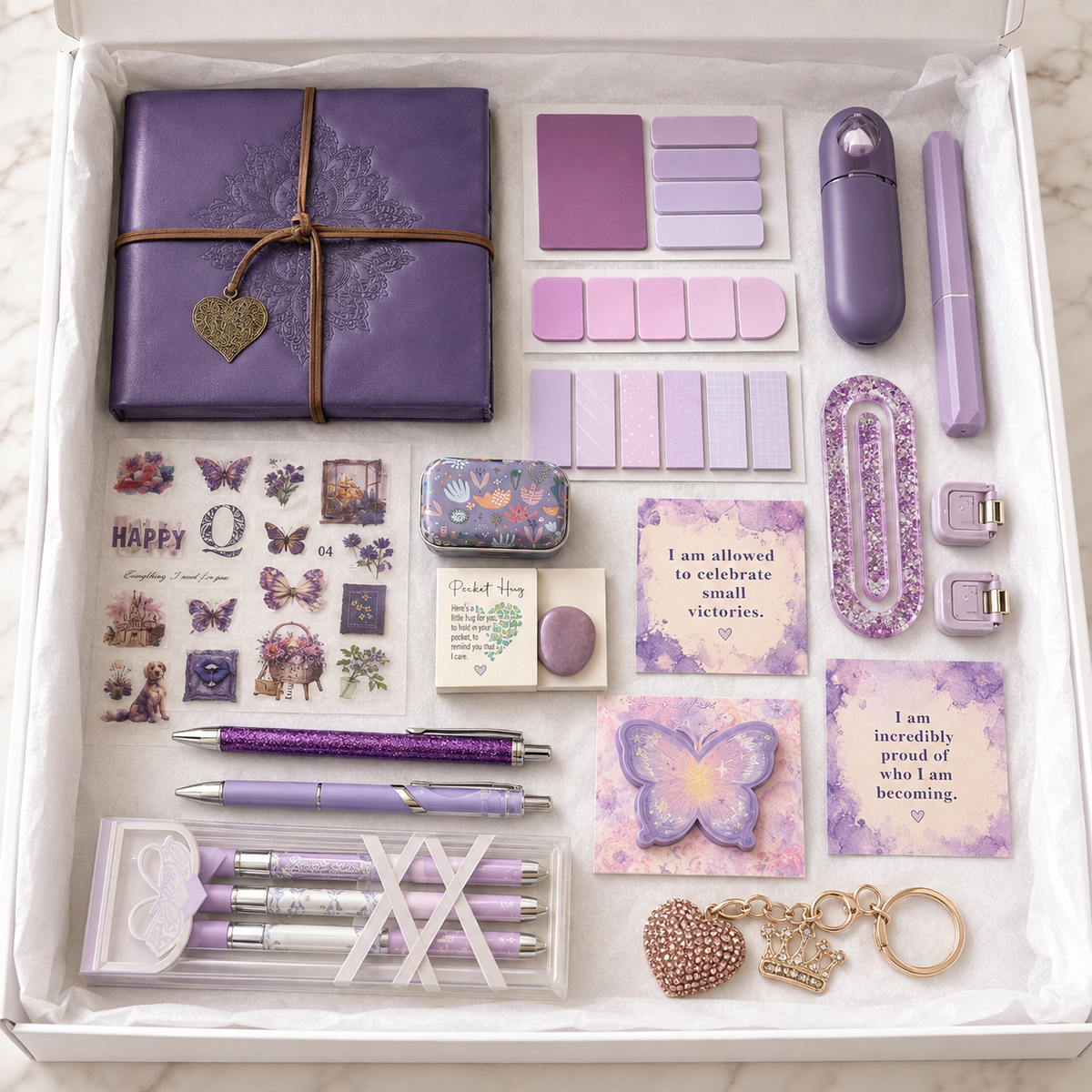 Lavender journal gift box in white box showing embossed mandala wrap journal with gold heart charm, boxed lavender-and-white pen set, purple glitter pen, lavender retractable pen, purple butterfly sticker sheet, page marker sets, butterfly sticky-note pad, affirmation cards, pocket hug keepsake, rose-gold rhinestone keychain, purple correction tape, floral accessory case, folding utility accessories and purple glitter-resin paperclip