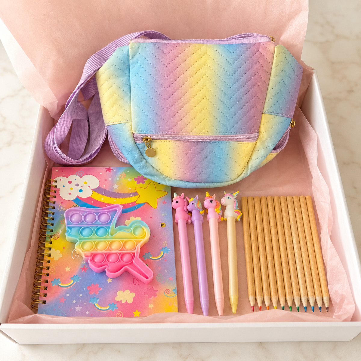 Pastel mini rainbow bag gift set in white box with blush-pink tissue paper showing quilted rainbow bag, spiral notebook, pop-it fidget toy, unicorn pens and colored pencils
