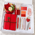 Red Journal and Diffuser Gift Box – true-red embossed mandala wrap journal, glossy red reed diffuser, red and cream rose bouquet, four decorative pens, red rhinestone heart keychain, silver feather bookmark, floral paperclip, and red page flags arranged in white gift box