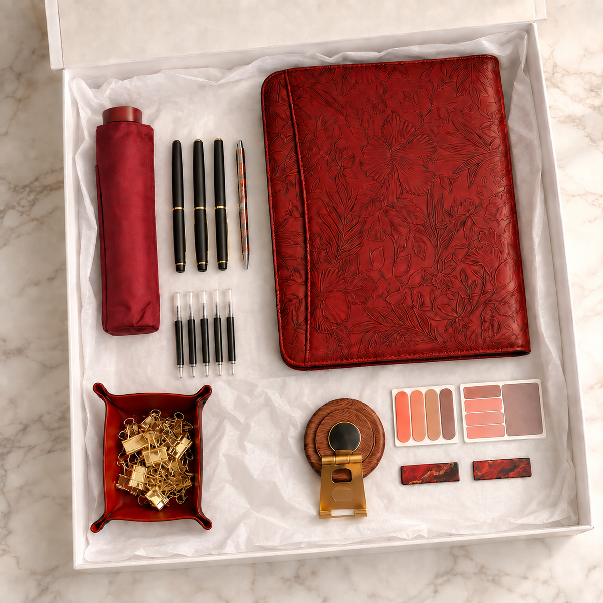 Burgundy executive gift box flatlay with floral embossed portfolio, travel umbrella, pen set, wooden phone stand, valet tray, and desk accessories