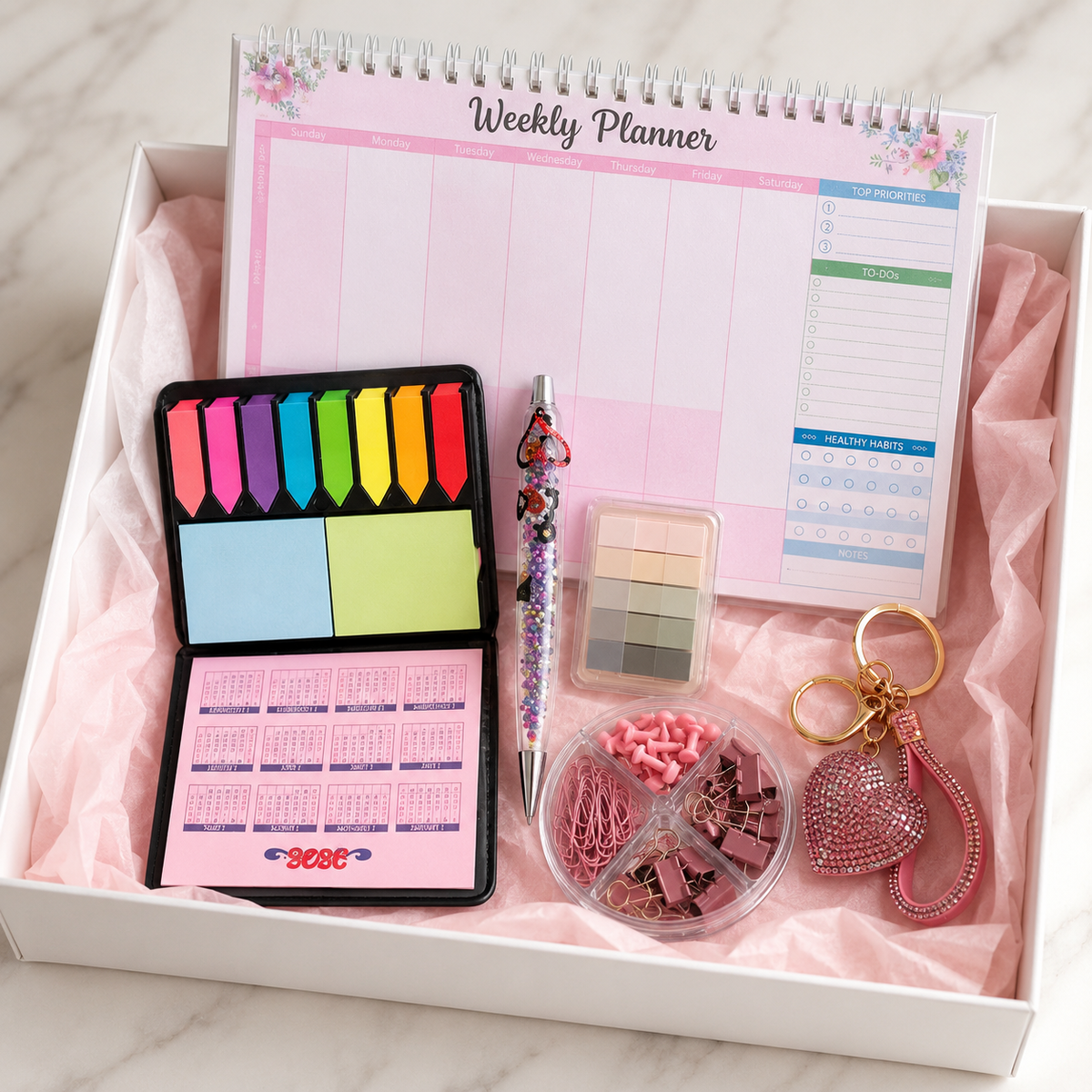 Pastel pink productivity gift box contents in white box with blush tissue paper showing weekly planner, beaded pen, sticky notes, page flags, desk organizer and rhinestone heart keychain