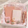 Pink everyday essentials gift box in white box with blush-pink tissue paper showing clear handled pouch, blush notebook, planner notepad, pastel page flags, pens, rhinestone keychain and badge holder